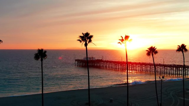 Cinematic aerial view of pier in Newport Beach, Orange County in California on sunny afternoon during golden hour sunset with palm trees in foreground.