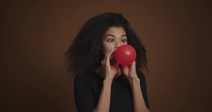 Portrait of african american girl inflates a red balloon at brown background