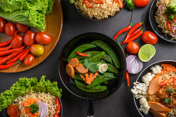 Stir-fried peas in a frying pan and noodles spicy in the bowl with ingredients on black cement background.
