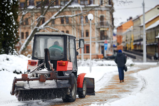 Tractor cleaning sidewalk from snow with snow plow and rotating brush. Municipal service removing snow, sprinkle salt and sand to prevent slipping. Pedestrian walking along cleared path on background