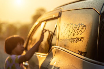Asian little girl writing or drawing heart symbol on wet mirror of her father SUV car in morning