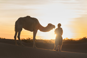 Fototapeta na wymiar Bedouin and camel on way through sandy desert Beautiful sunset with caravan on Sahara, Morocco Desert with camel and nomads Silhouette nomad man with dramatic sky Picturesque background nature concept