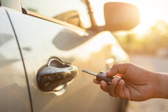 Hand holding remote car keys and press the button open or lock the silver car at outdoor parking lot with sunlight effect in morning or sunset time
