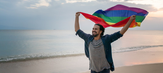 Cheerful guy with a rainbow flag on the beach. Young man holding a rainbow flag against the ocean...