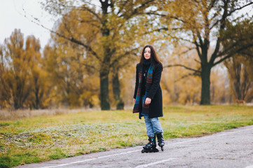 A pretty young girl in a coat and with a long scarf happily skates in the autumn park. Fighting the autumn depression. To overcome depression.