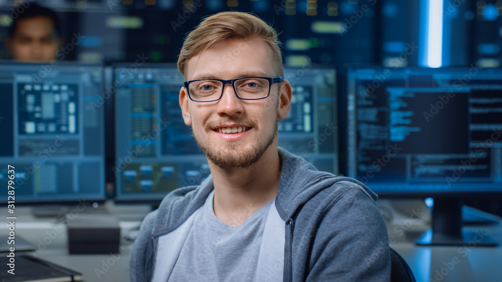 Poster Portrait of a Smart and Handsome IT Specialist Wearing Glasses Smiles, Behind Him Personal Computers with Screens Showing Software Program with Coding Language Interface in Data Center