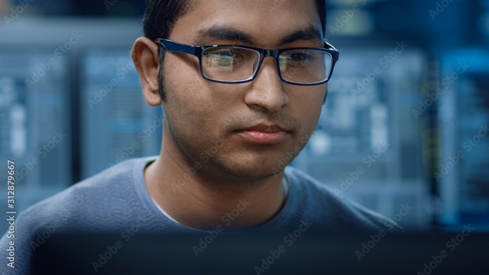 Poster Portrait of a Smart and Handsome Software Developer Wearing Glasses Working on a Personal Computer, Smiles at the Camera. In the Background Unfocused Personal Computers with Screens in Technical Room