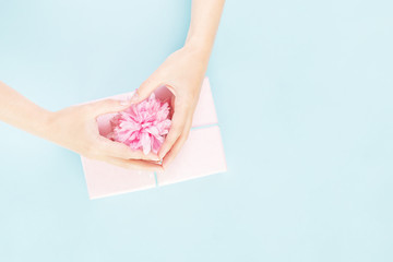 Female hands form a heart over pink gift box with festive flower on top against blue background. Present and celebration or giveaway for feminine bloggers. Mothers Valentines Women's Day. copy space