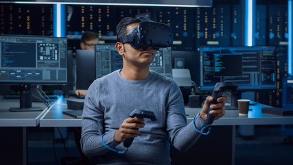 Portrait of Software Delevoper Wearing Virtual Reality Headset Using Controllers to Develop and Program VR Gaming Applications. In Background Technology Developing Studio with Computers and Monitors