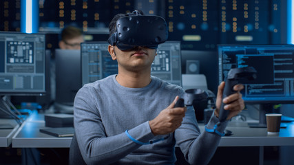 Portrait of Software Delevoper Wearing Virtual Reality Headset Using Controllers to Develop and Program VR Gaming Applications. In Background Technology Developing Studio with Computers and Monitors