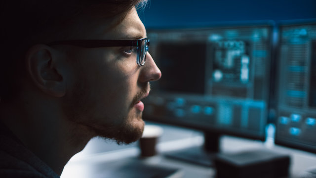 Portrait of IT Specialist Wearing Glasses Works on Personal Computer with Screens Showing Software Program with Coding Language Interface. Dark Colors. Close-up Shot