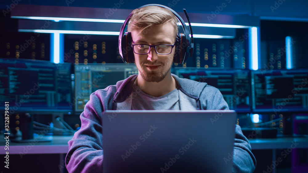 Poster Portrait of Software Developer / Hacker / Gamer Wearing Glasses and Headset Sitting at His Desk and Working / Playing on Laptop. In the Background Dark High Tech Environment with Multiple Displays.