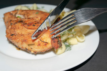 Chicken breast fried with spices, boiled pasta with herbs, metal fork and knife which cut a piece of the breast on a white plate.