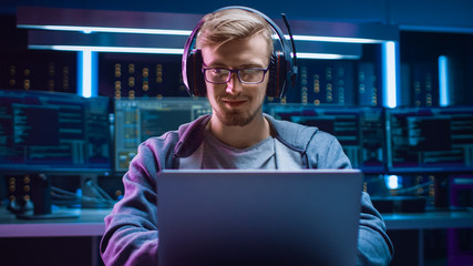 Portrait of Software Developer / Hacker / Gamer Wearing Glasses and Headset Sitting at His Desk and Working / Playing on Laptop. In the Background Dark High Tech Environment with Multiple Displays. - obrazy, fototapety, plakaty
