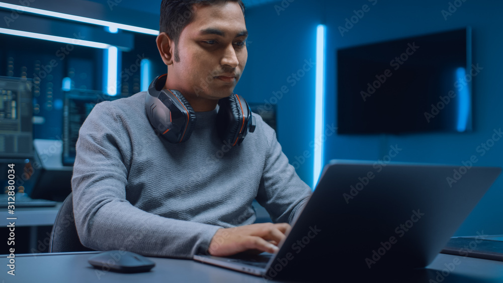 Poster Portrait of Software Developer / Hacker / Programmer Sitting at His Desk and Working on Laptop Computer In the Background Dark Neon High Tech Environment with Multiple Displays. 