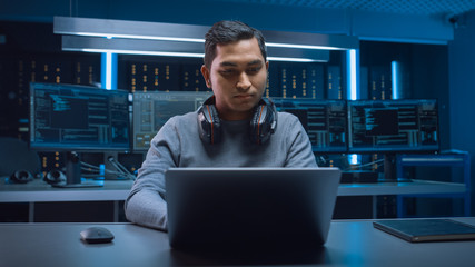 Portrait of Handsome Indian Software Developer / Hacker / Gamer Wearing Headset Sitting at His Desk and Working / Playing on Laptop. In Background Dark Neon High Tech Environment with Multiple Display