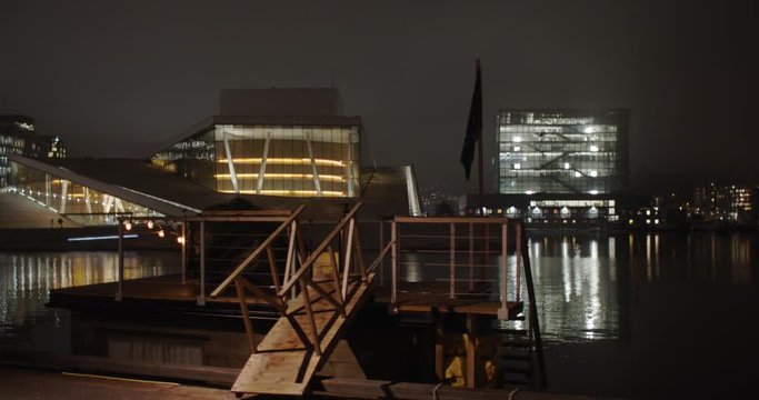 Cinematic wide angle 4K shot with parallax motion of sauna in front of illuminated Opera Music House and Edvard Munch gallery Lambda, with gold light reflecting in the fjord, at night in Oslo Norway.