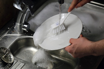 Doing dishes after cooking at home. Caucasian male doing the dishes. Closeup image. In this photo there is a white plate being washed with a lot of soup bubbles and running water. Color image.