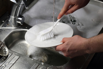Doing dishes after cooking at home. Caucasian male doing the dishes. Closeup image. In this photo there is a white plate being washed with a lot of soup bubbles and running water. Color image.