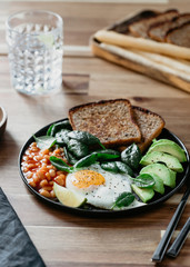 Healthy breakfast or lunch at home or cafe with fried egg, avocado, toasts, beans and fresh spinach on a wooden table.