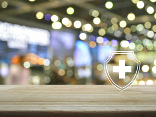 Cross shape with shield flat icon on wooden table over blur light and shadow of shopping mall, Business healthy and medical care insurance concept