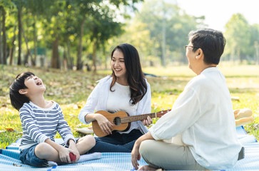 Asian family having picnic in fresh green park together. Mother playing Ukelele guitar with smile while grandmother looking at child singing song and laughing with happiness. Relax family activity.