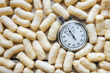 A close-up of a round glass vintage stopwatch against the background of yellow corn sticks. Time, hours, lunch break. Composition and concept. Food for speed.