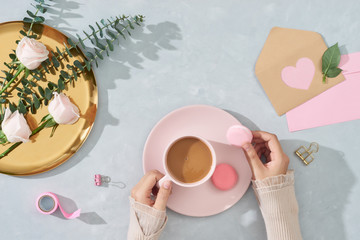 Woman hands holding macaroon and coffee cup with flowers and card on light blue background. Top view, flatlay