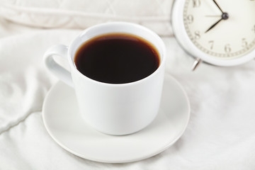 Cup of coffee in white cup with white alarm clock on white bed. Close up.