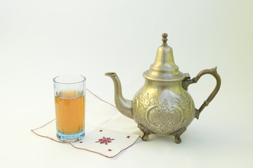 Old and worn traditional Moroccan teapot with glass of mint tea on embroidered handkerchief and on a white background close up