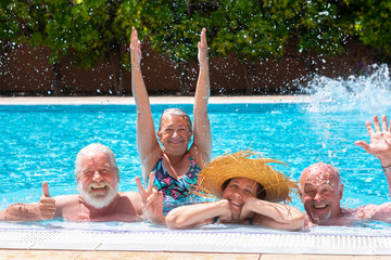 Cheerful group of four senior people floating in outdoor swimming pool raising splashes of water. They smile relaxed on vacation under the bright sun