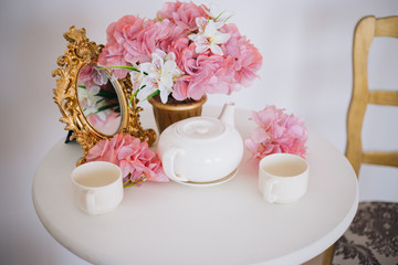 Tea table, mugs and a teapot white teapot with a pink bouquet of flowers in the rococo style. Golden vintage mirror. White pink eltion interior