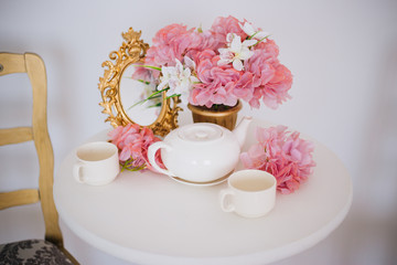 Tea table, mugs and a teapot white teapot with a pink bouquet of flowers in the rococo style. Golden vintage mirror. White pink eltion interior