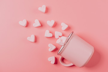 a pink mug lies on its side against a pink background, light-pink marshmallows sprinkled from it in the form of hearts