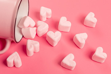 a pink mug lies on its side against a pink background, light-pink marshmallows sprinkled from it in the form of hearts