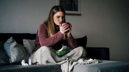 nachdenkliche junge Frau ist krank und sitz auf Sofa und trinkt Tee