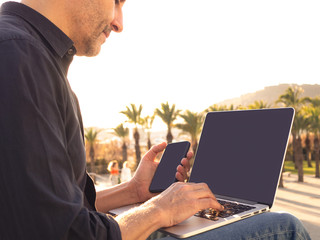 Man working remotely on phone and laptop sitting outside at sunset