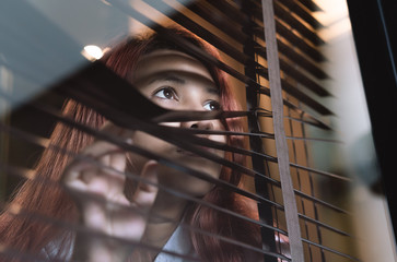 Young woman indoors looking through window blinds during quarantine