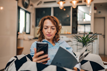 Fototapeta premium Portrait of a woman reading a text message on smart phone while reading a book at home.