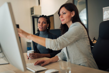 Manager instructing trainee, pointing at computer at modern workplace.