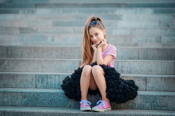 A beautiful girl in a fluffy black skirt tutu and pink sneakers sits on the steps.