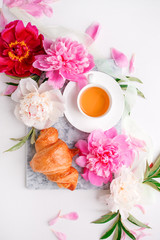 Obraz na płótnie Canvas Breakfast for Valentines day with cup of tea, peony flowers and croissant on gray murble table and white background from above, flat lay, wedding, birthday, romance concept