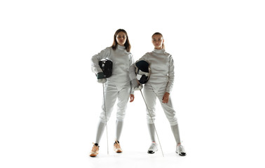 Teen girls in fencing costumes with swords in hands isolated on white studio background. Young female models training, posing confident. Copyspace. Sport, youth, healthy lifestyle, motion, action.