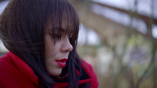 portrait of a brunette with bright lipstick and piercings on her face. she's wearing a red jacket with the collar turned up. her fringe fall on her eyes. close up