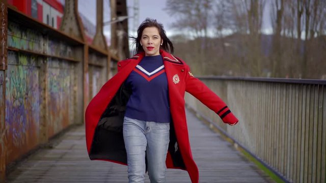 a brunette with bright lipstick and a piercing on her face is walking on a railway bridge. she's wearing a blue jumper and a red coat. the woman raises her hands. passing train