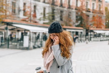 Fototapeta premium Young blonde woman with winter clothes speaks on the mobile phone while carrying a cup of coffee in the other hand
