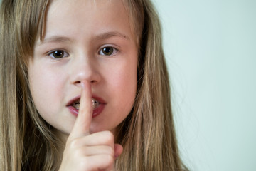Fototapeta premium Close-up portrait of little child girl with long hairholding point finger to her lips making hush gesture.