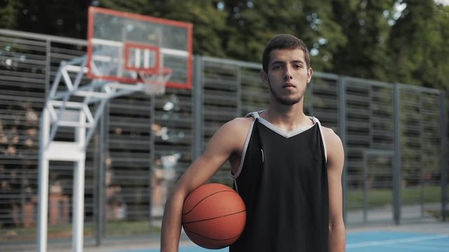 Close Up Portrait of Young Slim Fit Caucasian Basketball Player in Black Singlet Holding Ball Standing at Street Basketbal Court and Looking to Camera. Healthy Lifestyle and Sport Concept.