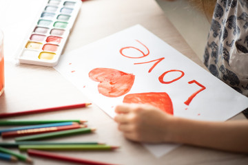 child paints with watercolors watercolor red heart on a landscape sheet on a wooden table