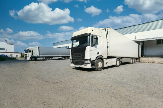 White Truck With A White Trailer At Unloading At The Warehouse.Warehouse Complex With An Asphalt Pad And Truck With Blue Sky